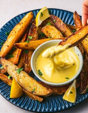 a potato wedge being dipped into a bowl of aioli that's on a plate with more potato wedges and lemons.
