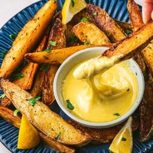 A potato wedge being dipped into the bowl of aioli that's resting on a plate with more potato wedges and lemons.