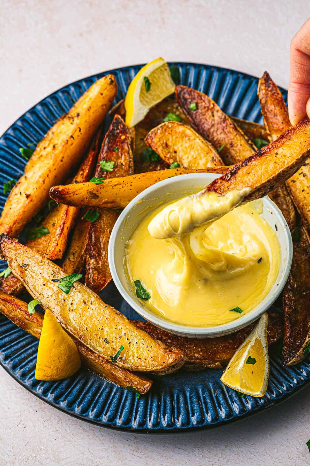 a potato wedge being dipped into a bowl of garlic aioli that's on a plate with more potato wedges and lemons.
