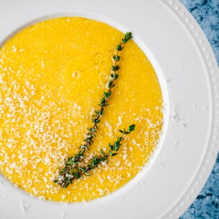 close up of a bowl of polenta topped with parmigiano reggiano cheese and a sprig of thyme.