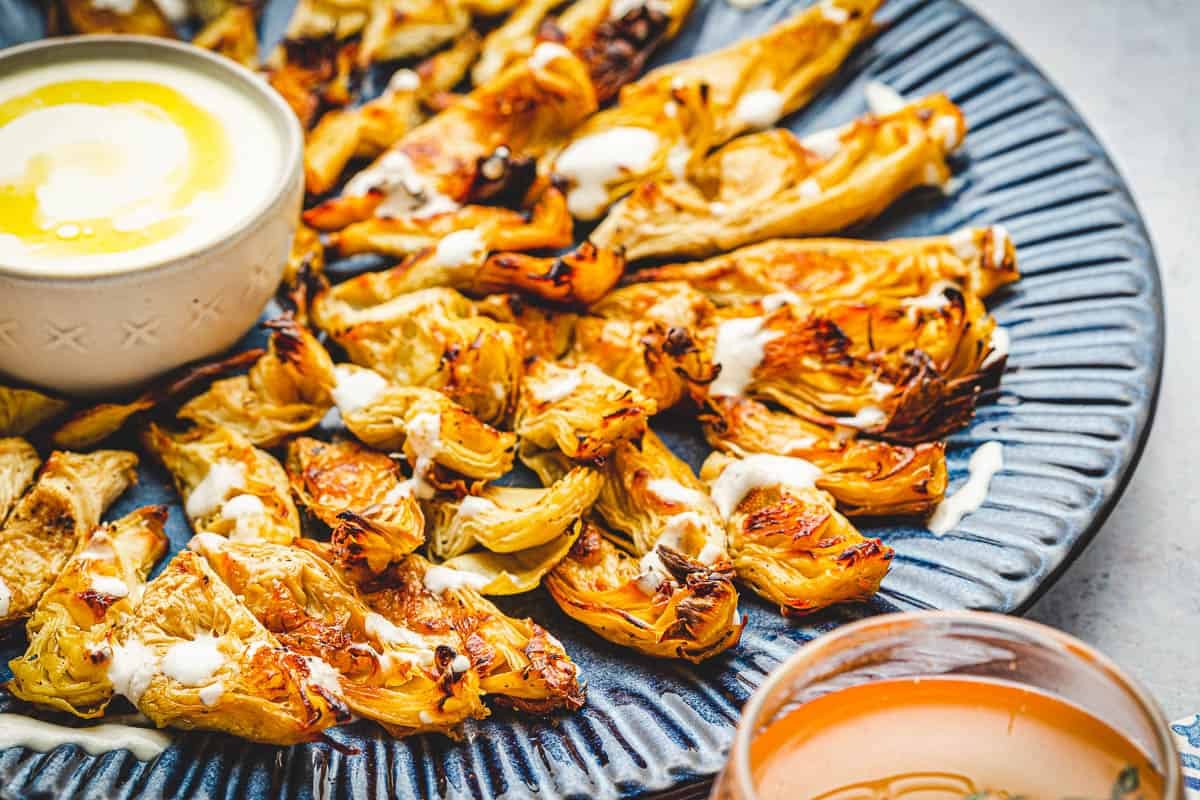 Side shot of roasted artichoke hearts drizzled with feta dressing and a white bowl of feta dressing on the side.