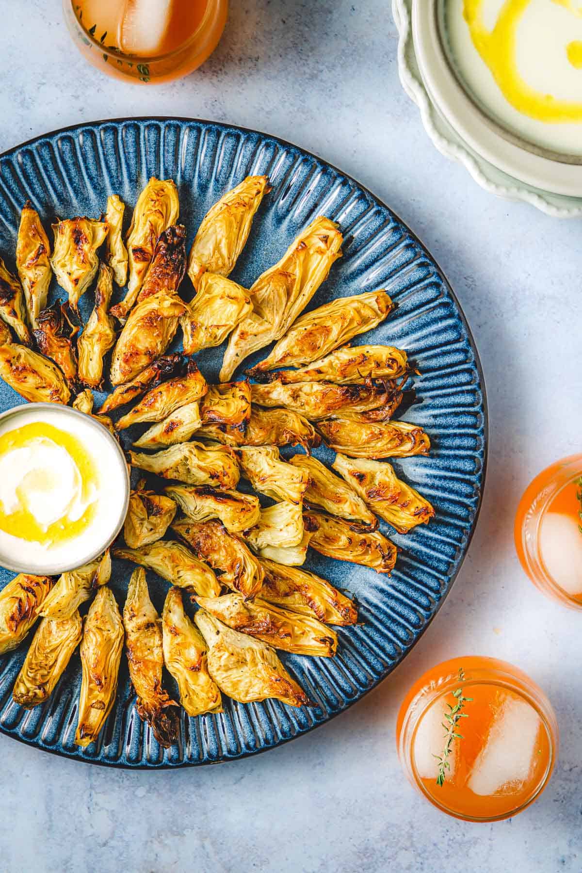 Overhead shot of golden brown roasted artichoke hearts drizzled with creamy feta dressing on a blue plate with three cocktails on the side and a white marble background.