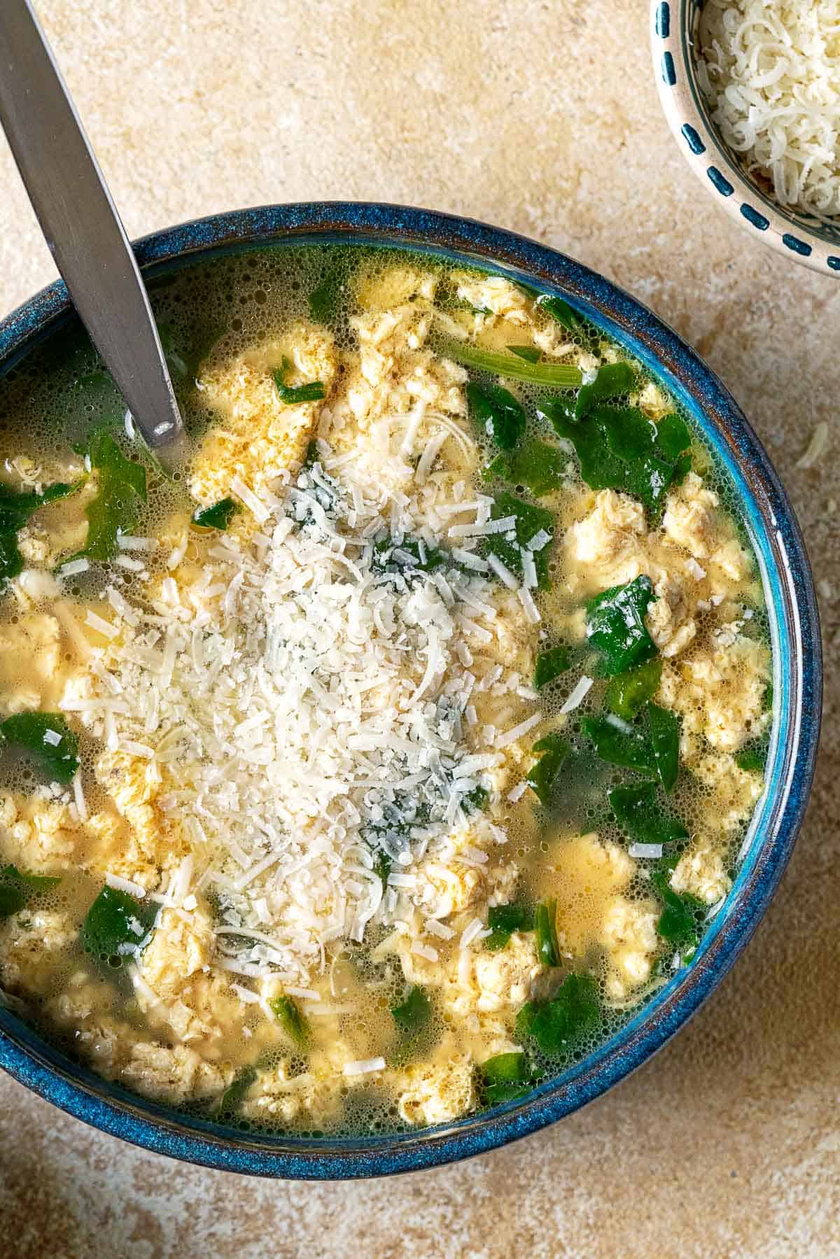 Overhead shot of a blue bowl with Stracciatella Soup with wilted spinach and freshly grated Parmesan cheese.