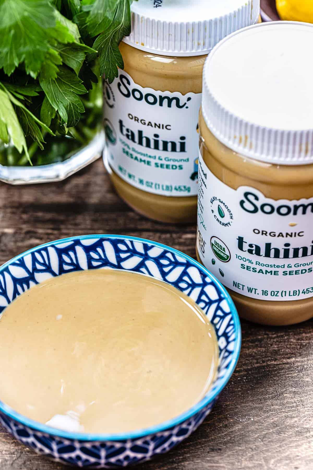 Bowl of tahini with two jars of tahini and fresh parsley in the background.