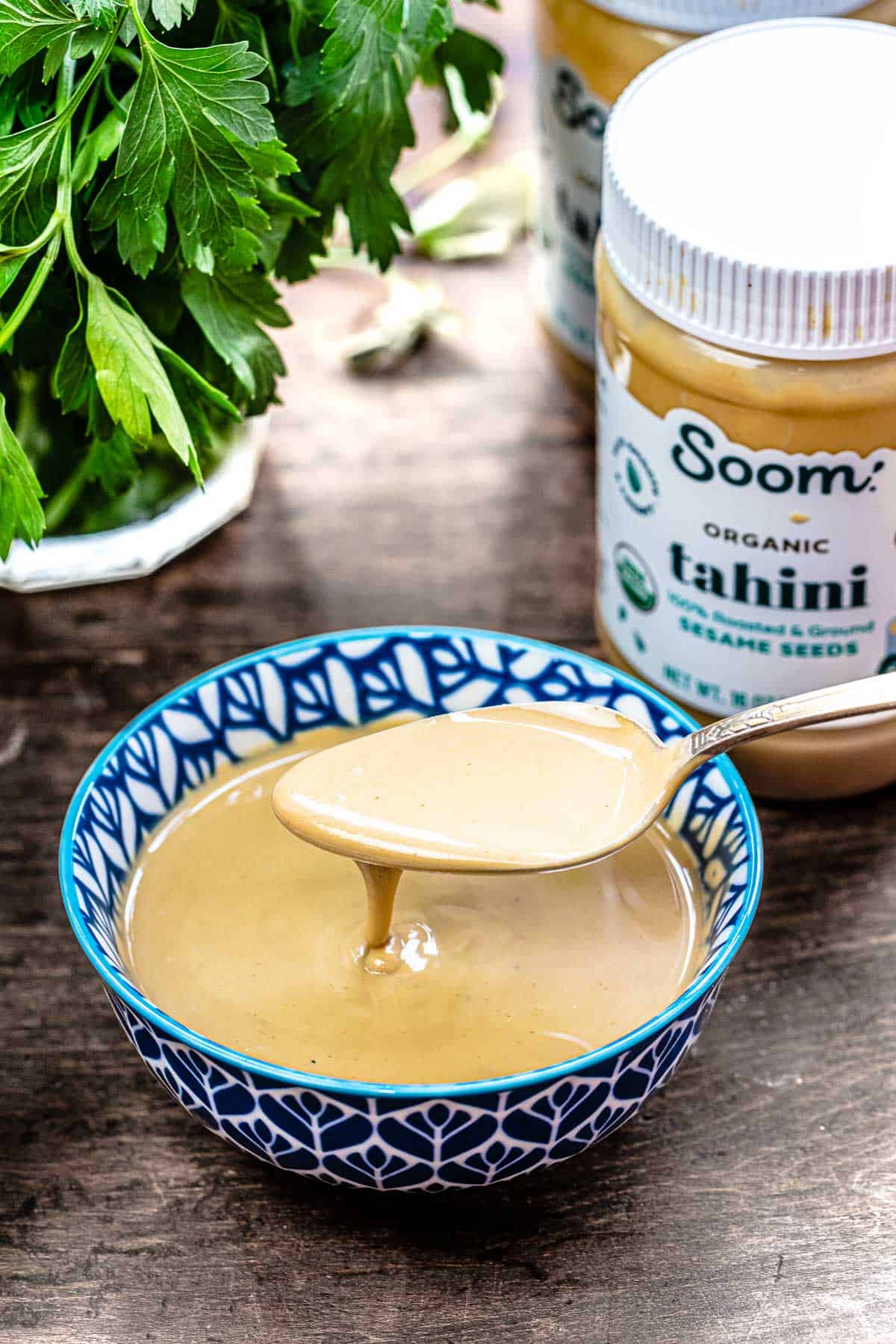 A spoon lifting tahini from the bowl as it drips down with fresh parsley in the background.