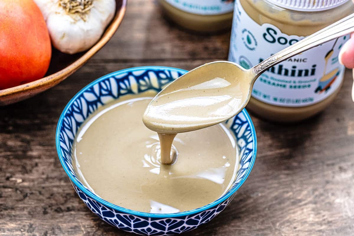 A spoon dripping with tahini over a bowl with vegetables and a jar of tahini in the background.