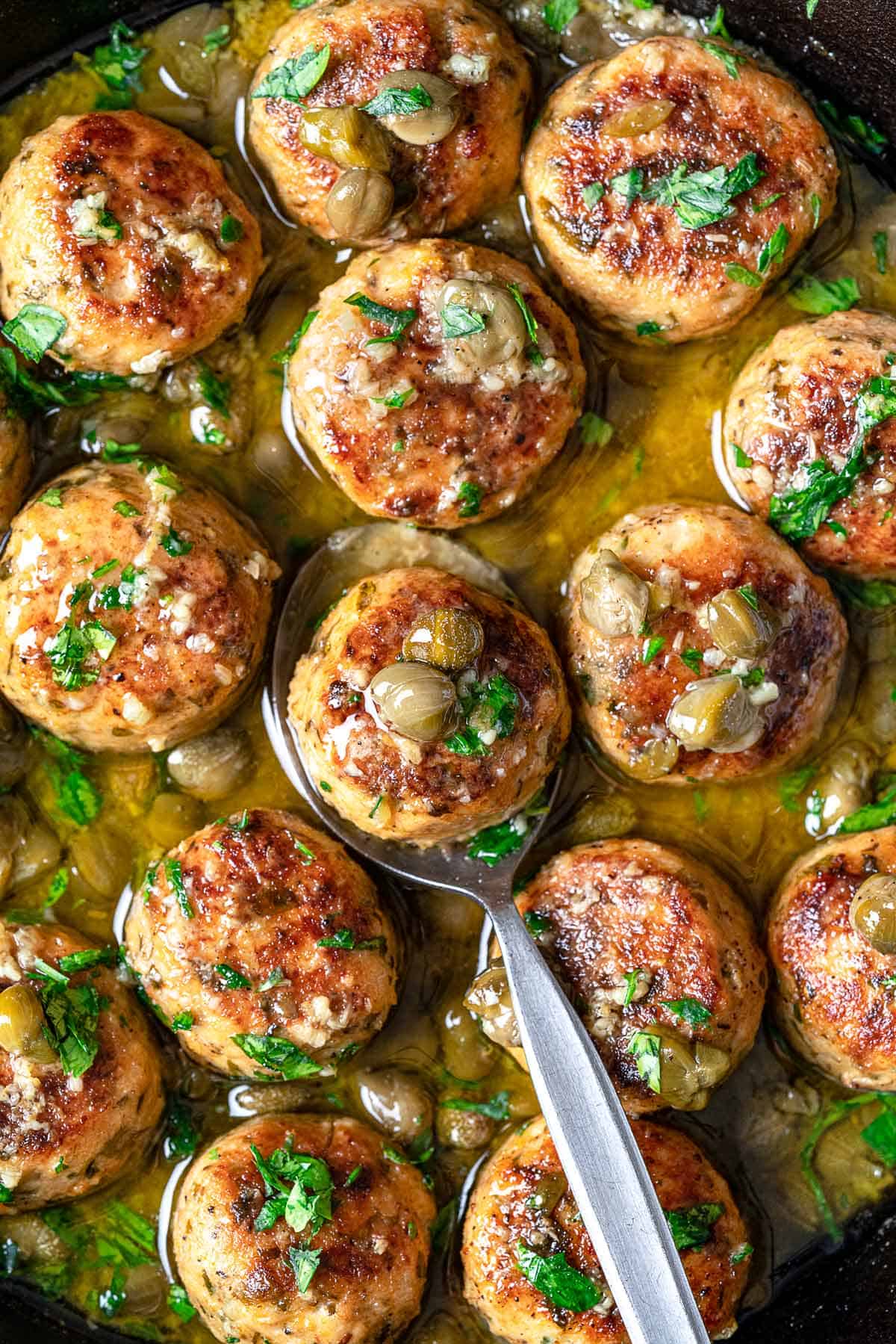 one chicken piccata meatball being lifted out of a skillet-full of chicken piccata meatballs with a spoon.