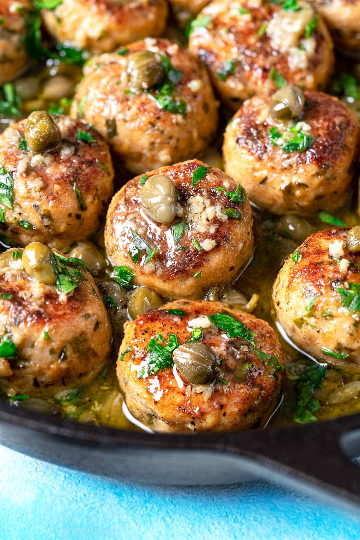 close up of chicken piccata meatballs in a cast iron skilet topped with parsley and capers.