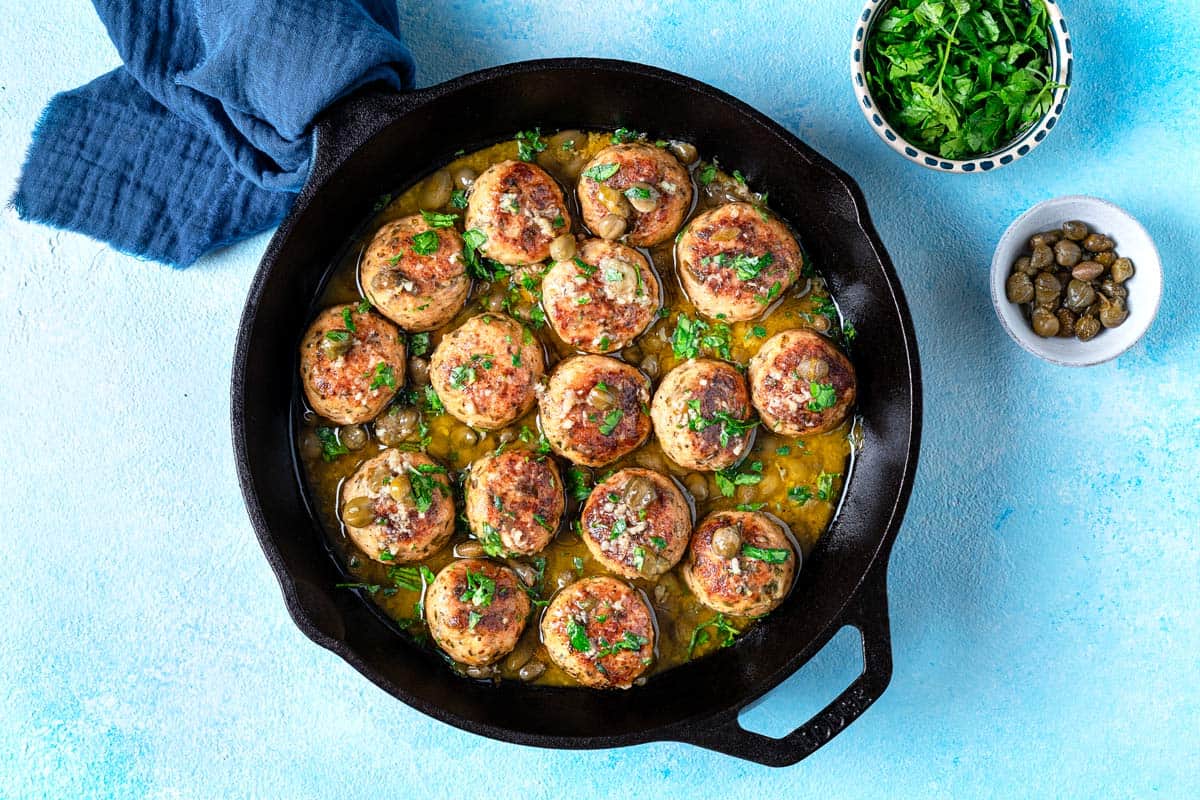 chicken piccata meatballs in a cast iron skillet next to a bowl of chopped parsley and a bowl of capers.