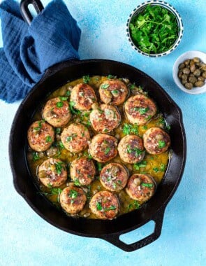 chicken piccata meatballs in a cast iron skillet next to a bowl of chopped parsley and a bowl of capers.