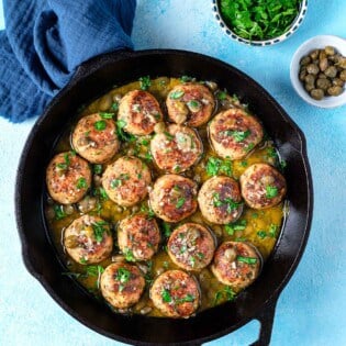 chicken piccata meatballs in a cast iron skillet next to a bowl of chopped parsley and a bowl of capers.