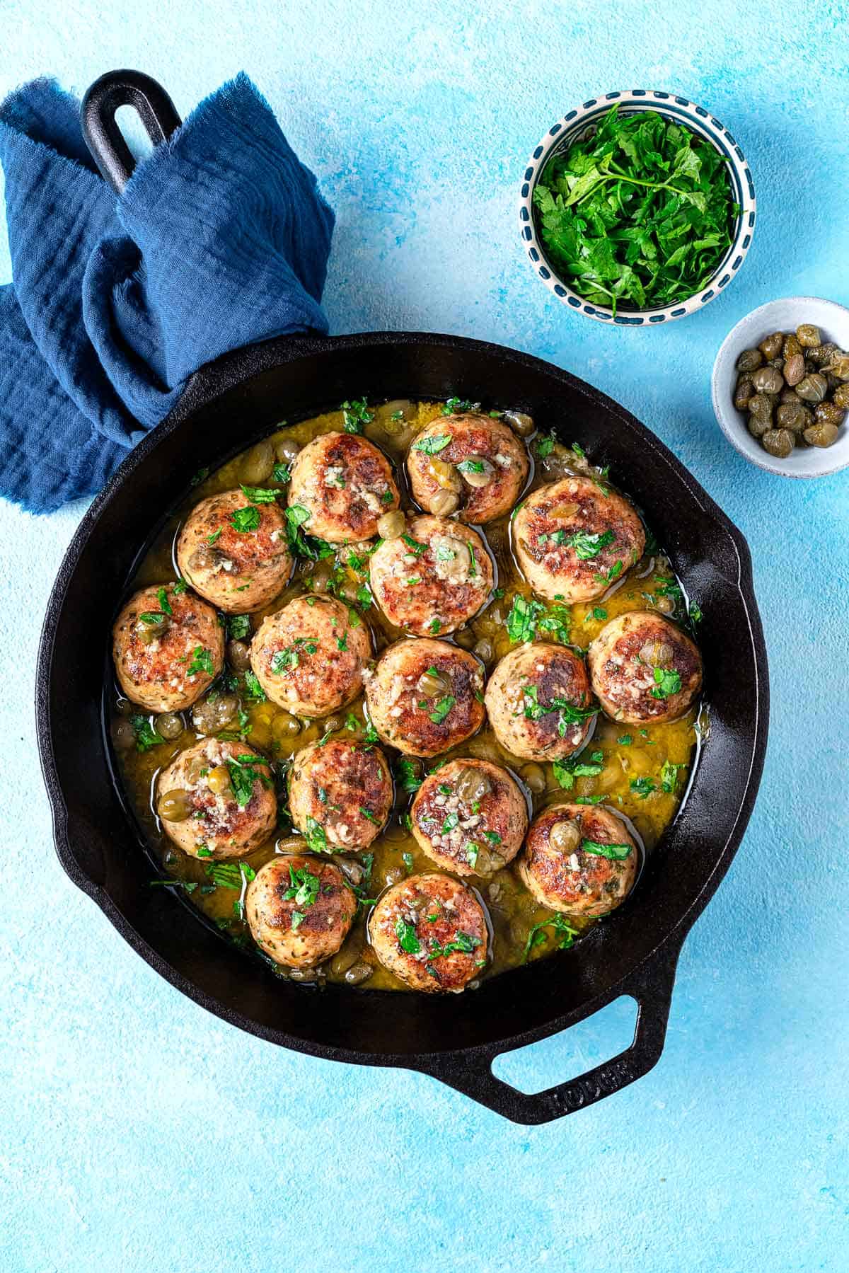 chicken piccata meatballs in a cast iron skillet next to a bowl of chopped parsley and a bowl of capers.