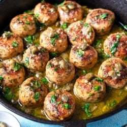 chicken piccata meatballs in a cast iron skillet next to a bowl of chopped parsley and a bowl of capers.