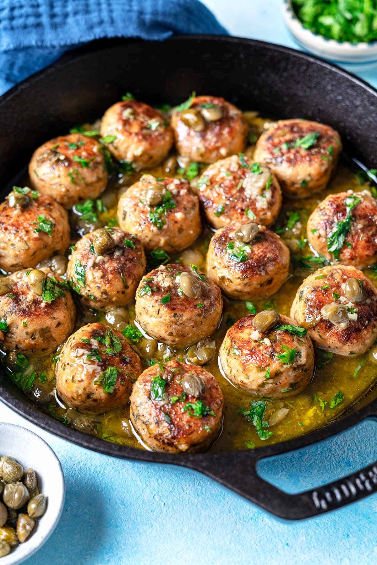 chicken piccata meatballs in a cast iron skillet next to a bowl of chopped parsley and a bowl of capers.