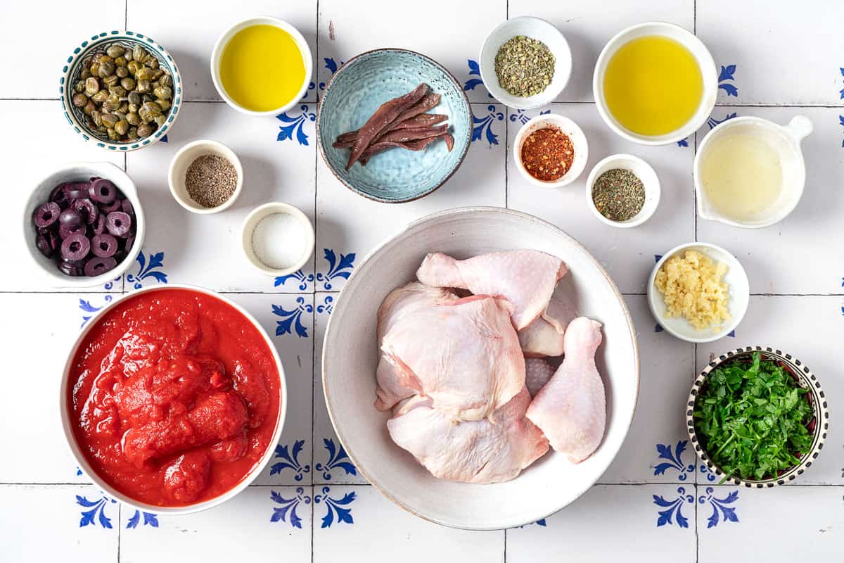 ingredients for chicken puttanesca including pieces of bone-in skin-on chicken, salt, pepper, olive oil, italian seasoning, lemon juice, anchovy fillets, minced garlic, whole peeled tomatoes, kalamata olives, capers, red pepper flakes, oregano, and chopped parsley.