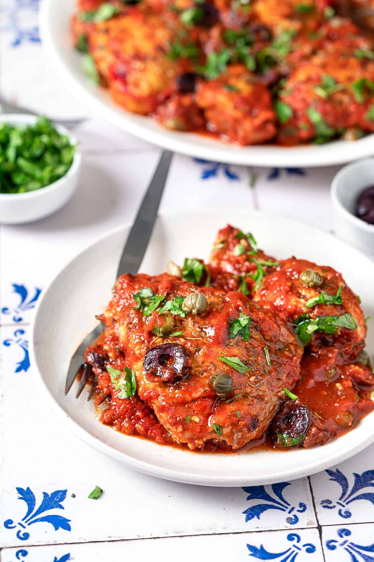 two pieces of chicken puttanesca on a plate with a fork in front of a serving platter of chicken puttanesca and a bowl of chopped parsley.