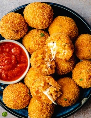 two arancini italian fried risotto balls opened up on a pile of other arancini balls on a plate with a bowl of marinara.