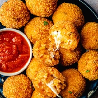 two arancini italian fried risotto balls opened up on a pile of other arancini balls on a plate with a bowl of marinara.