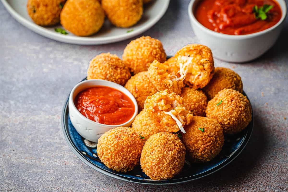 arancini italian fried risotto balls on a plate with a bowl of marinara with another bowl of marinara and plate of arancini in the background.