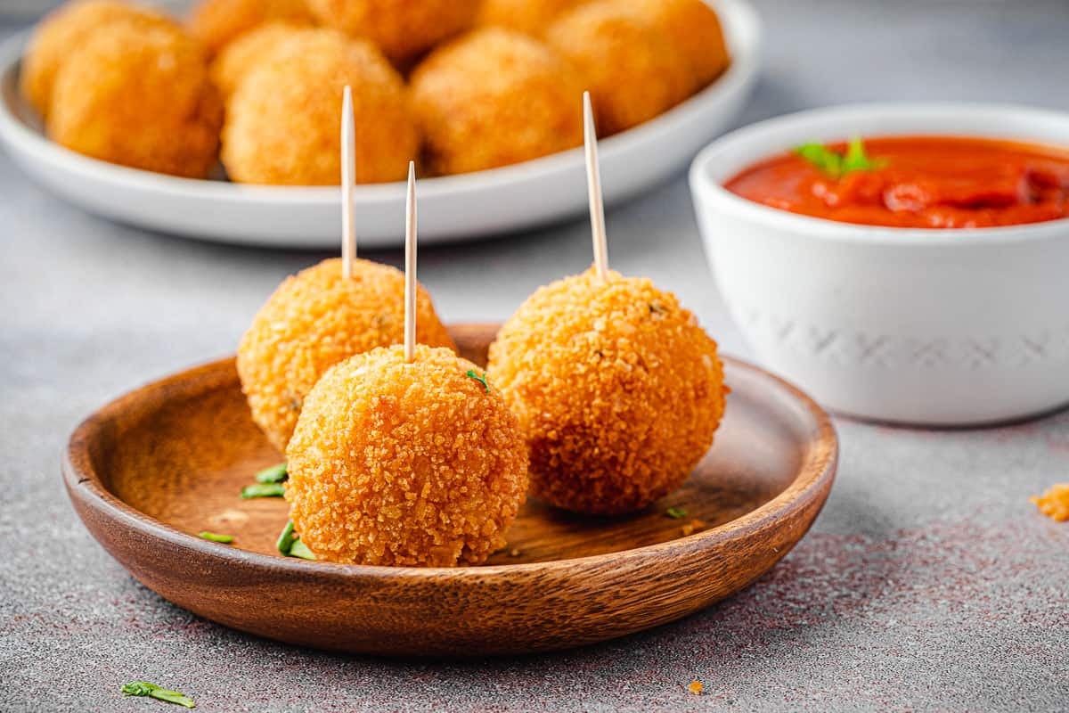 3 arancini italian fried risotto balls on a plate with toothpick in each one in front of a bowl of marinara and another plate of arancini.