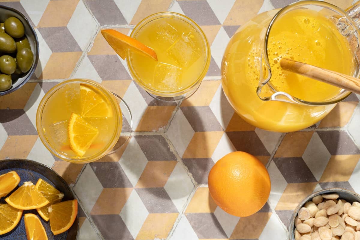 overhead shot of a pitcher of agua de valencia, two glasses of agua de valencia, an orange, a bowl of almonds, a plate of orange slices and a bowl of green olives.