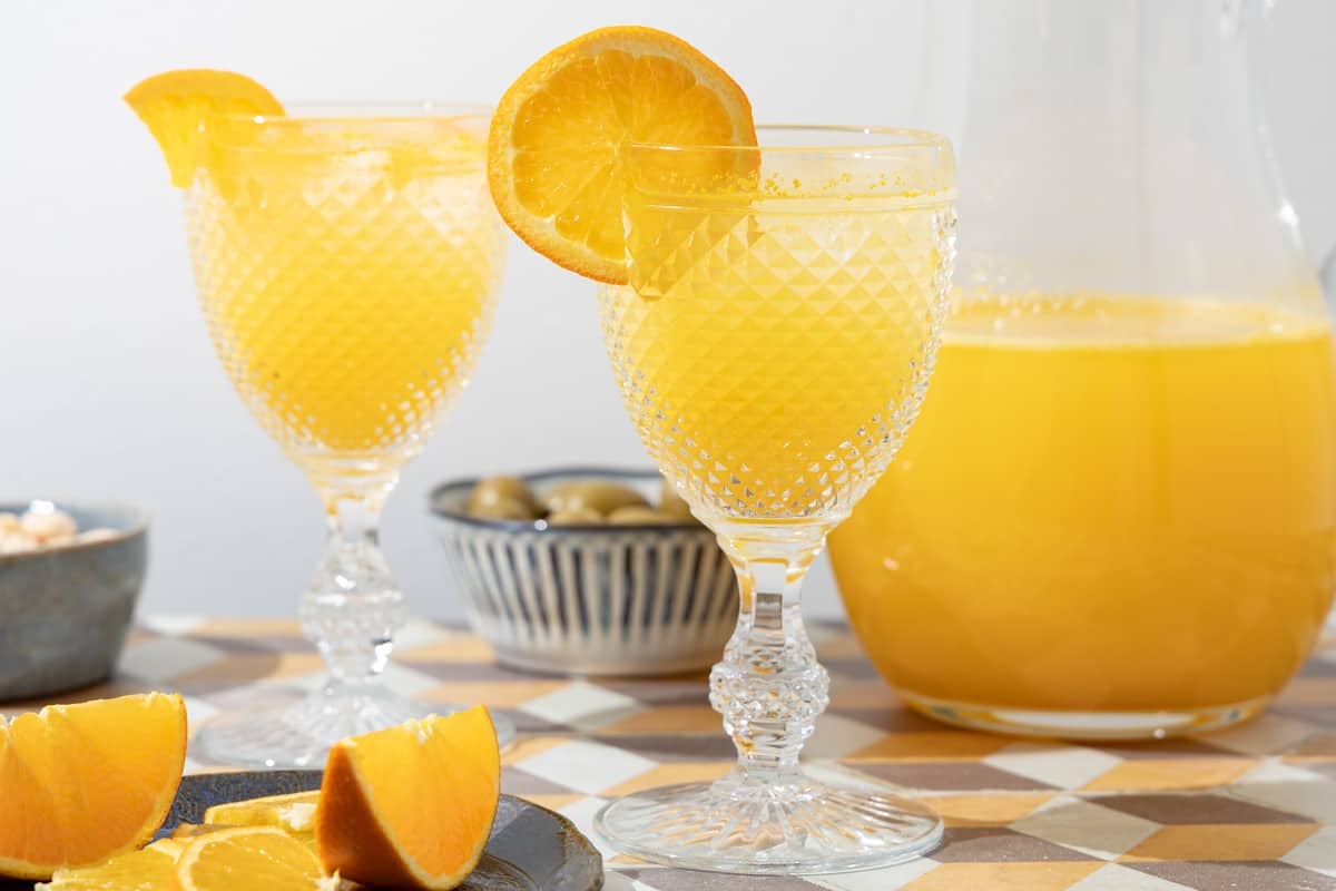 two glasses of agua de valencia garnished with orange slices in front of a pitcher of agua de valencia.