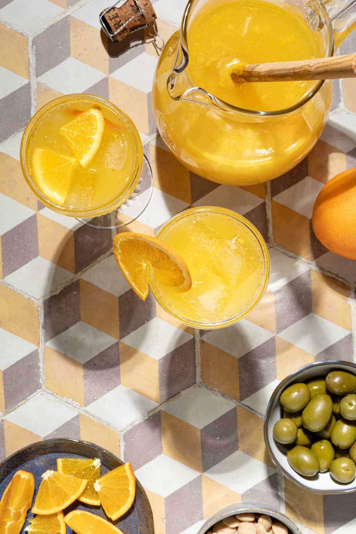 overhead shot of a pitcher of agua de valencia, two glasses of agua de valencia, an orange, a bowl of almonds, a plate of orange slices and a bowl of green olives.