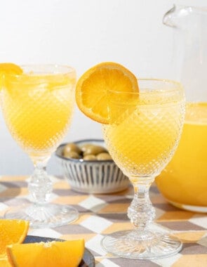 two glasses of agua de valencia garnished with orange slices in front of a pitcher of agua de valencia.