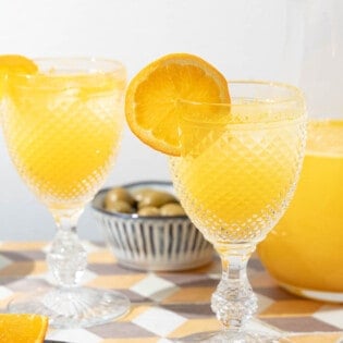 two glasses of agua de valencia garnished with orange slices in front of a pitcher of agua de valencia.