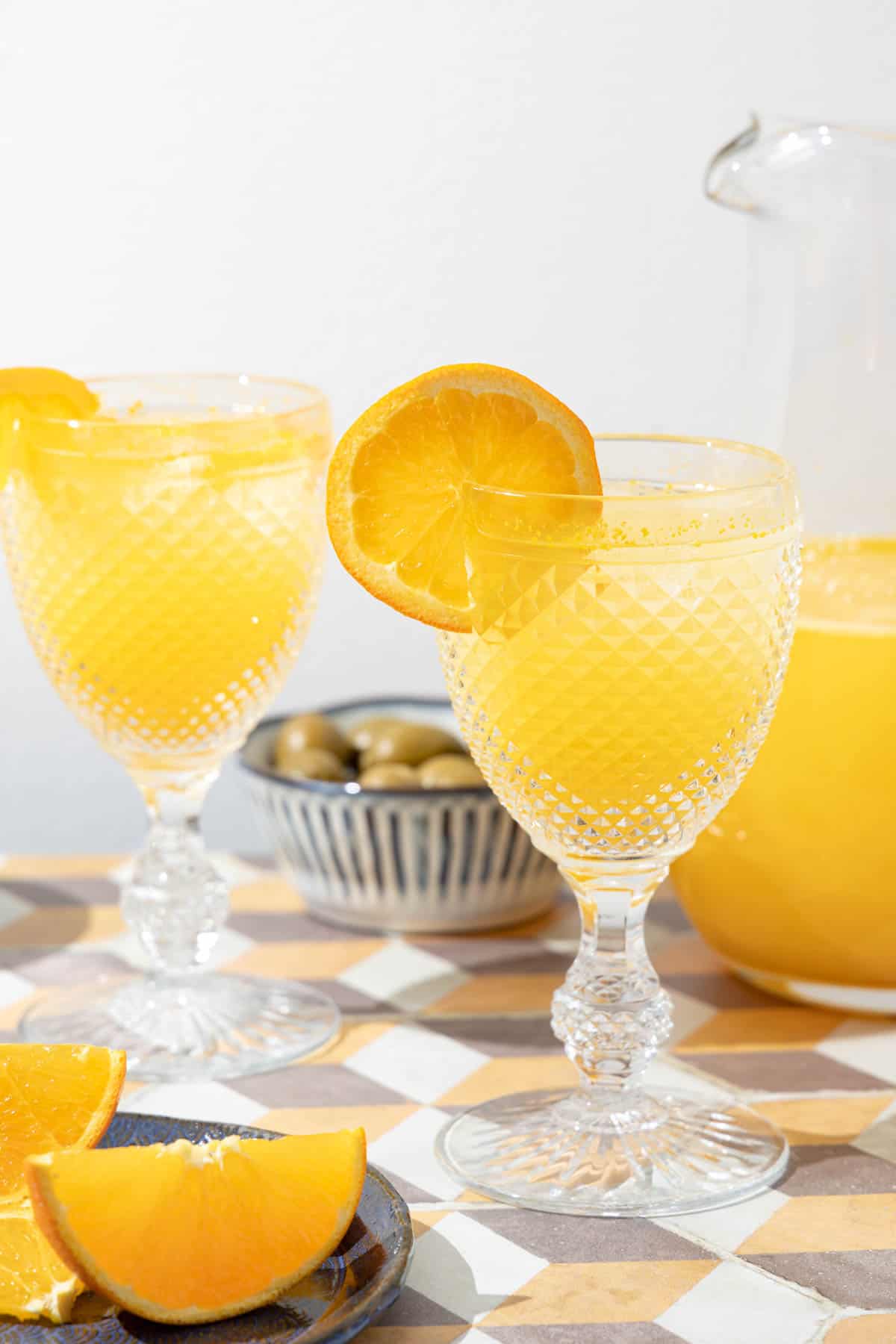 two glasses of agua de valencia garnished with orange slices in front of a pitcher of agua de valencia.