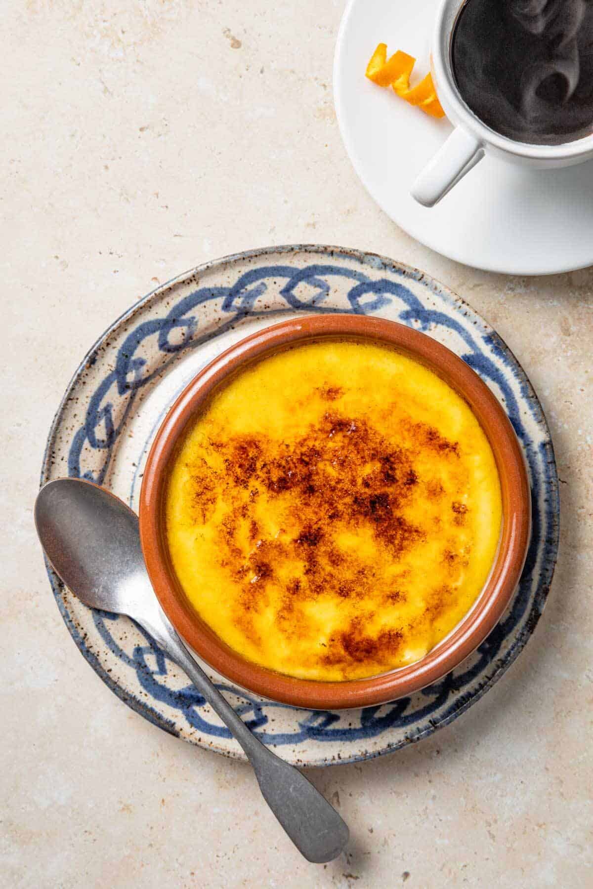 Overhead shot of Crema Catalana with a silver spoon, coffee, and an orange zest twist on the side.