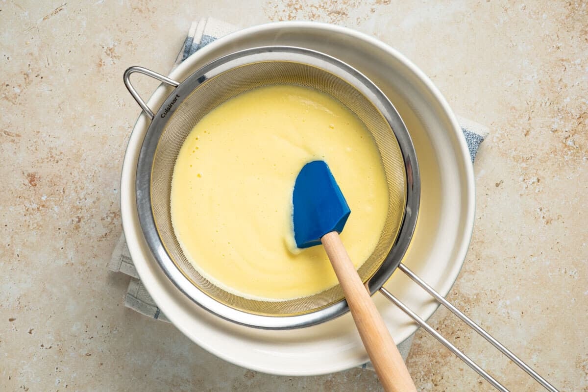 Crema Catalana being pushed through a fine mesh sieve over a white bowl with a blue rubber spatula.