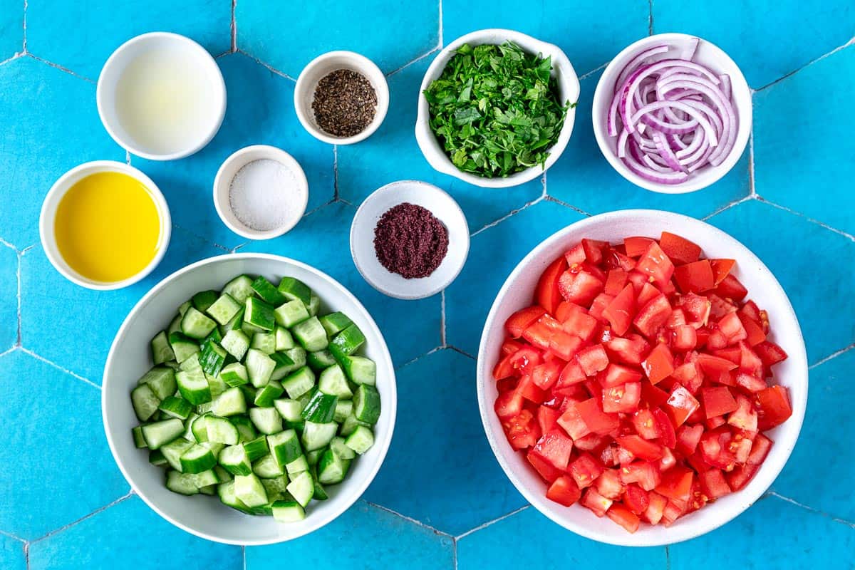 ingredients for cucumber tomato salad including cucumber, tomatoes, parsley, red onion, salt, pepper, sumac, olive oil and lemon juice.
