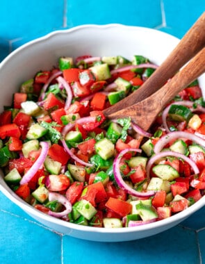 cucumber tomato salad in a bowl with wooden serving utensils next to a bowl of chopped parsley and lemons.