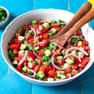 cucumber tomato salad in a bowl with wooden serving utensils next to a bowl of chopped parsley and lemons.