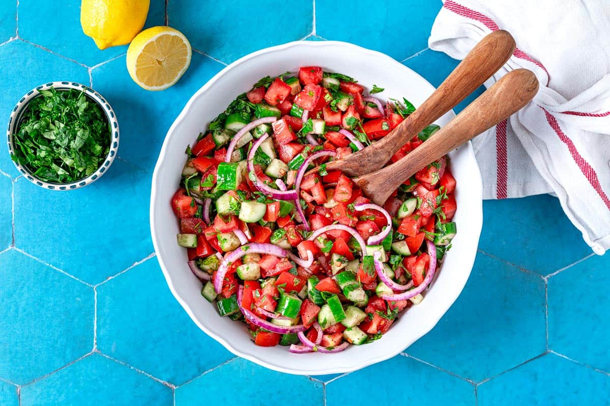 cucumber tomato salad in a bowl with wooden serving utensils next to a bowl of chopped parsley, lemons and a kitchen towel.
