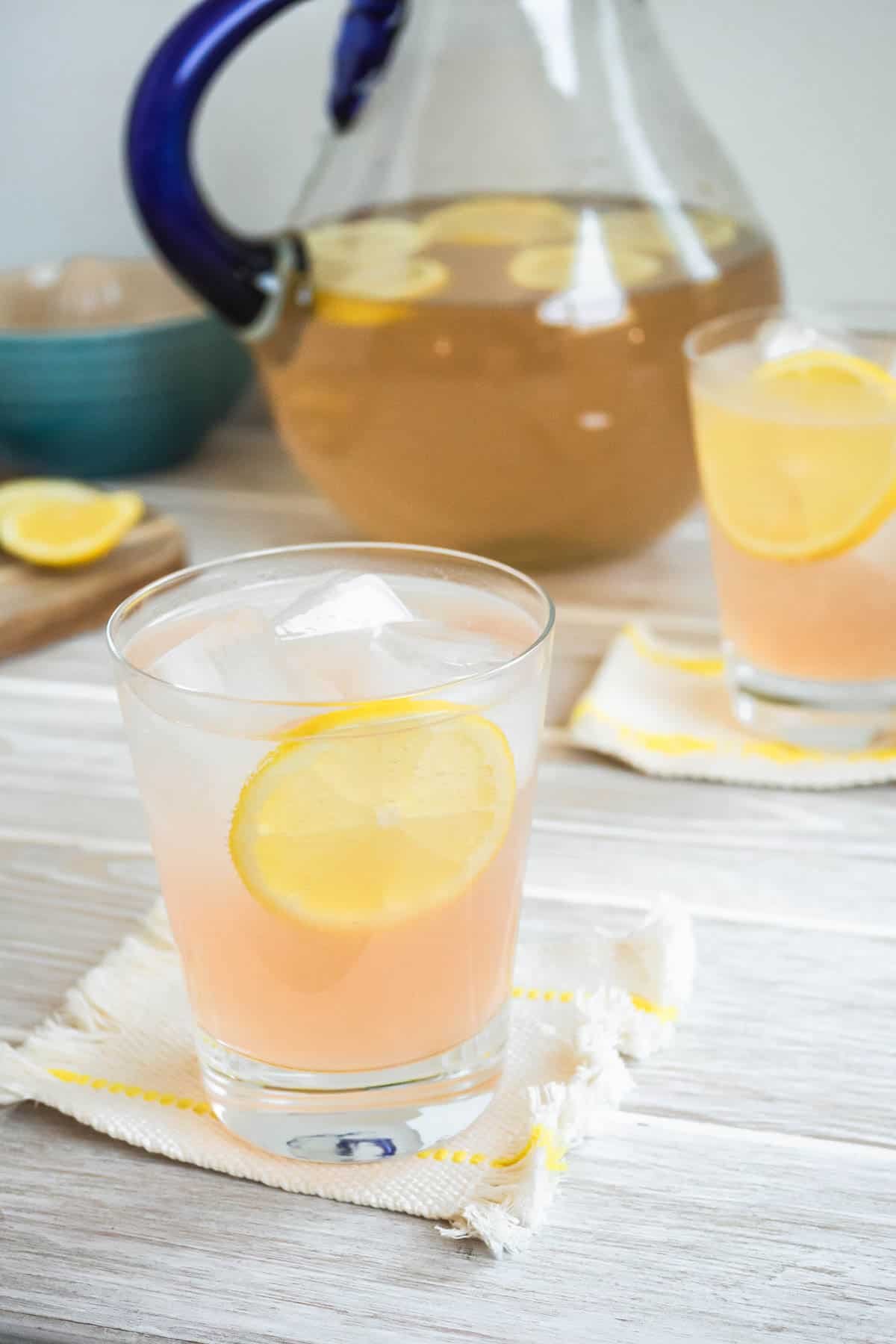 a glass of rose lemonade on a coaster, garnished with a lemon wheel, with another glass and pitcher of rose lemonade in the background.