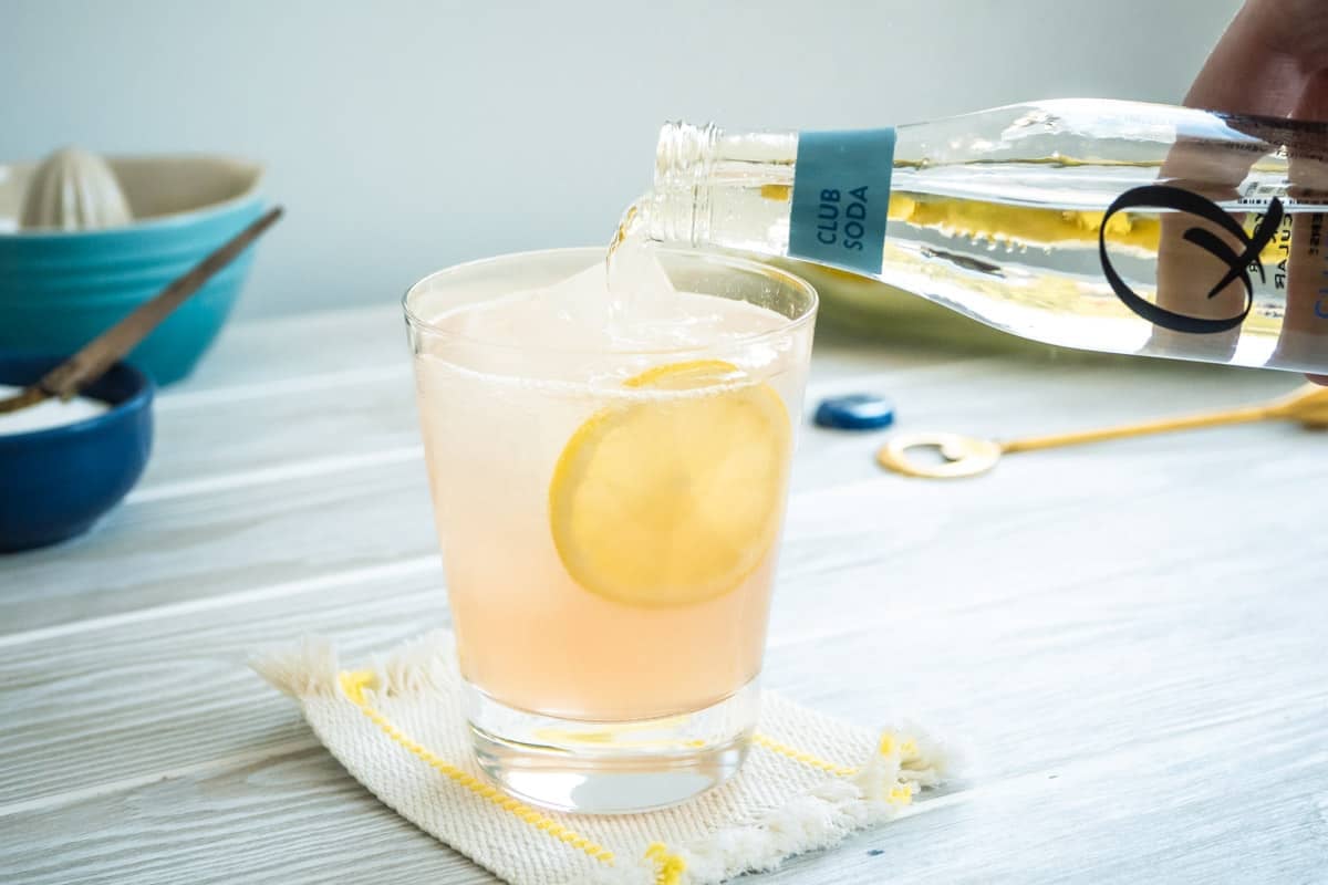 club soda being added to a glass of rose lemonade on a coaster garnished with a lemon wheel.