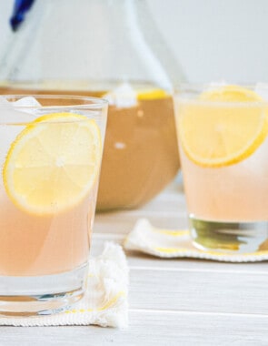 two glasses of rose lemonade on coasters, each garnished with a lemon wheel, with a pitcher of rose lemonade in the background.