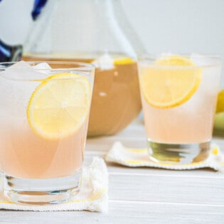 two glasses of rose lemonade on coasters, each garnished with a lemon wheel, with a pitcher of rose lemonade in the background.