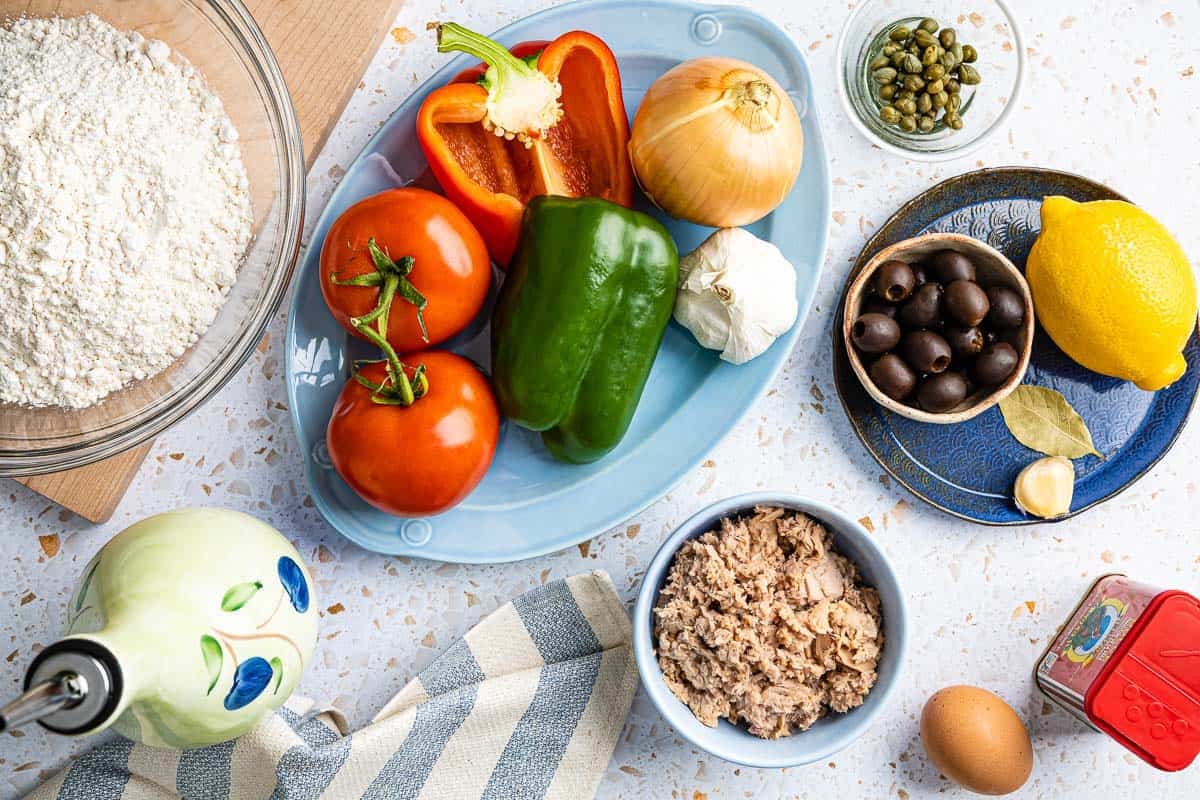 ingredients for tuna empanadillas including flour, olive oil, tomatoes, bell peppers, onion, garlic, capers, black olives, lemon, bay leaf, an egg and paprika.