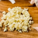 Pile of minced garlic on a wooden cutting board with one unpeeled garlic clove on the side.