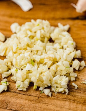 Pile of minced garlic on a wooden cutting board with one unpeeled garlic clove on the side.