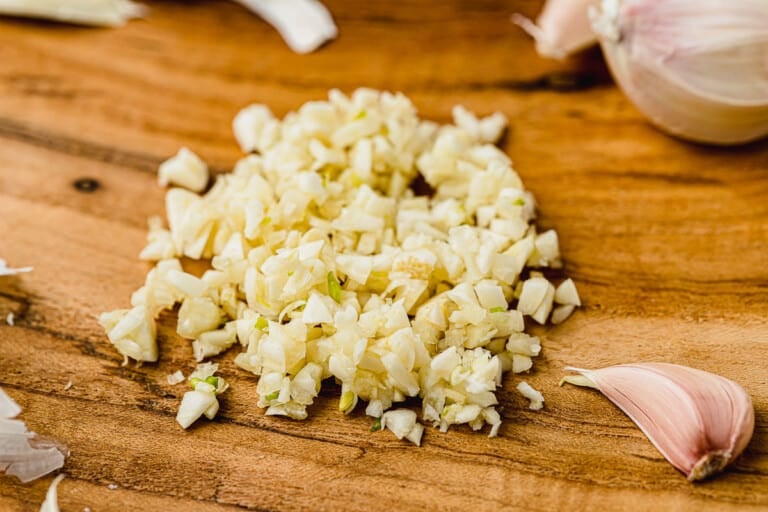 Pile of minced garlic on a wooden cutting board with one unpeeled garlic clove on the side.