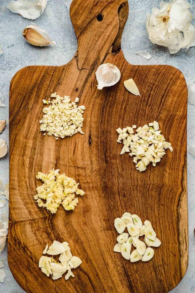 Wooden cutting board showing minced, chopped, grated, smashed, and sliced garlic.