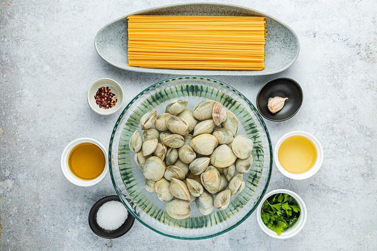 Ingredients for linguini with clams, including unshucked clams, garlic, oil, parsley, salt, red pepper flakes, white wine, and uncooked linguini.