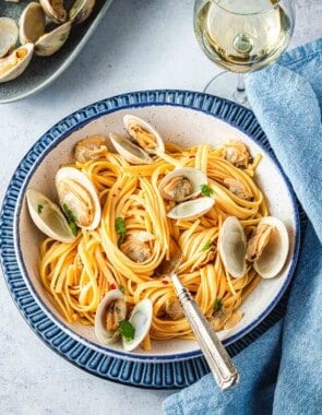 A serving of linguini with clams with a blue linen napkin and glass of white wine on the side.