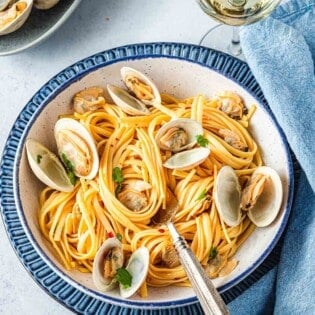 A serving of linguini with clams with a blue linen napkin and glass of white wine on the side.