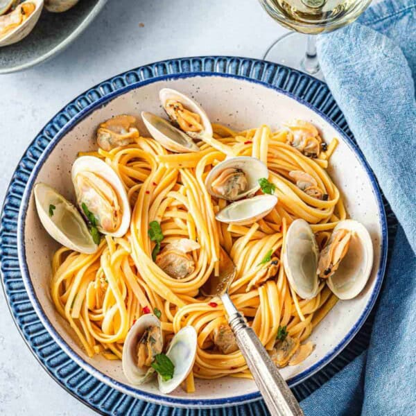 A serving of linguini with clams with a blue linen napkin and glass of white wine on the side.