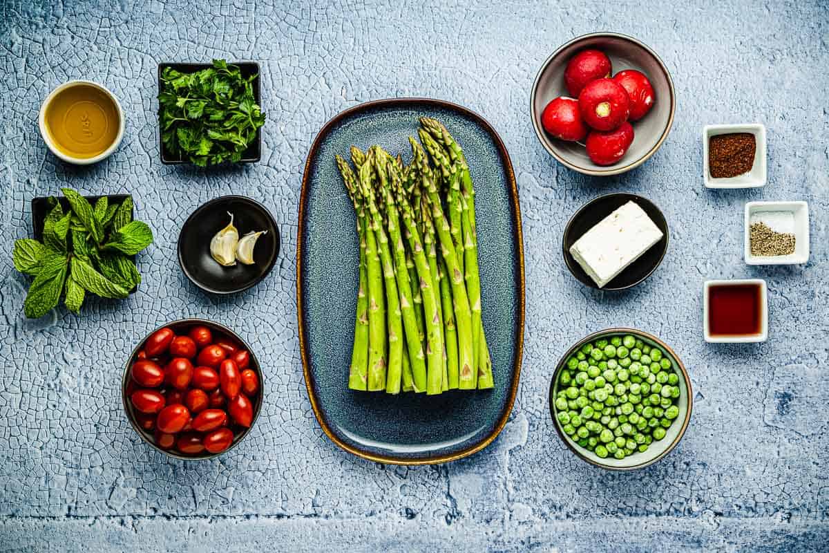 Ingredients for Asparagus Salad, including asparagus, pea, cherry tomatoes, radishes, mint, parsley, feta, red wine vinegar, garlic, salt, pepper, sumac, and oil.